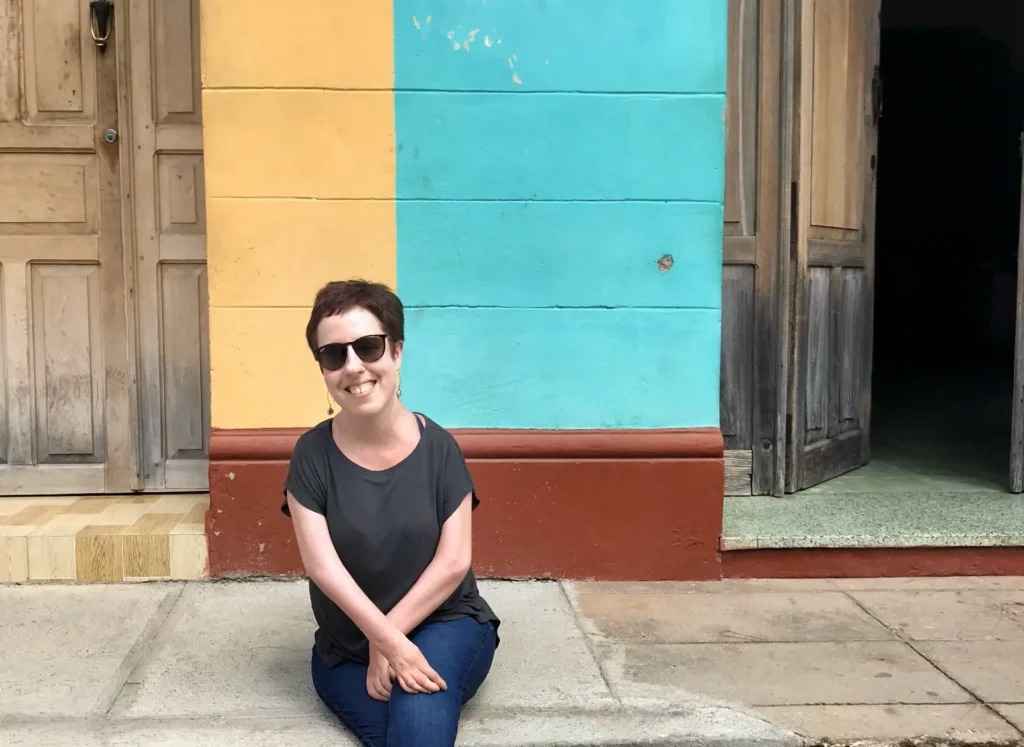 Woman in sunglasses sits smiling on a ledge, in front of a vibrant yellow and turquoise wall with wooden doors, evoking a cheerful, relaxed vibe.
