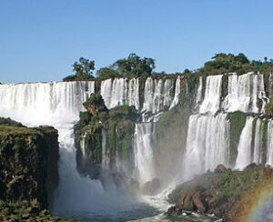 A majestic waterfall cascades over rocky cliffs, surrounded by lush greenery under a clear blue sky. A faint rainbow appears in the mist below.