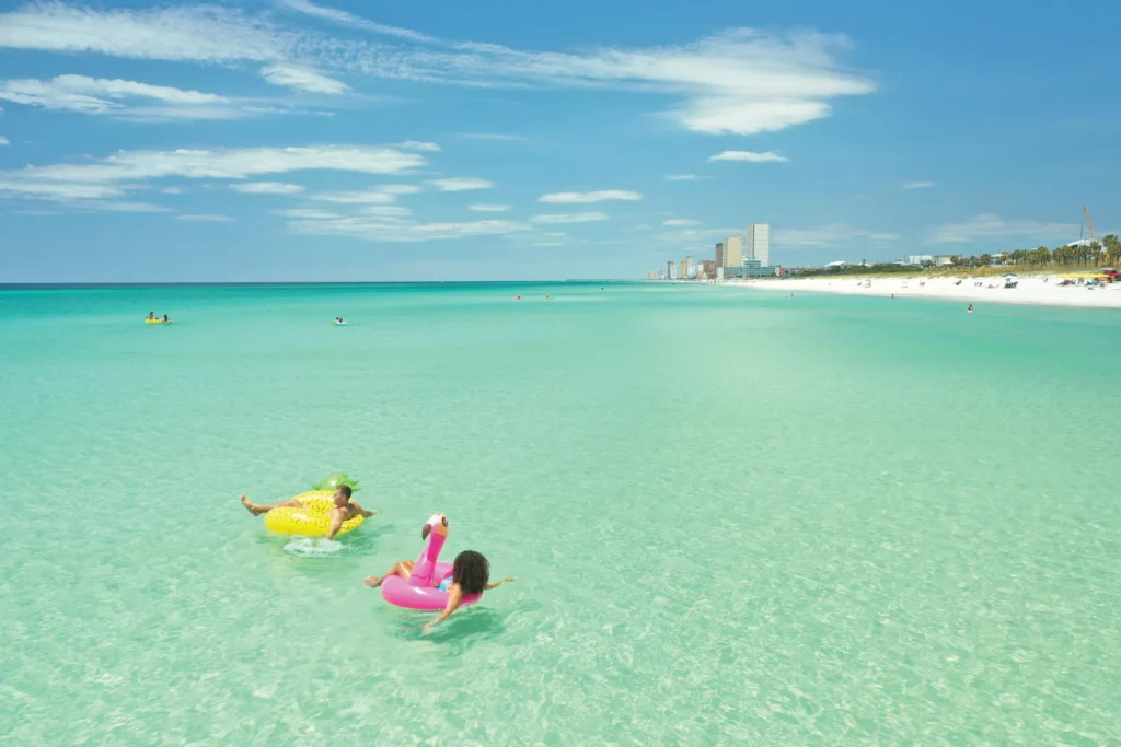 Two people float on colorful inflatable rings in a crystal-clear ocean, with a sunny beach and skyline in the background.