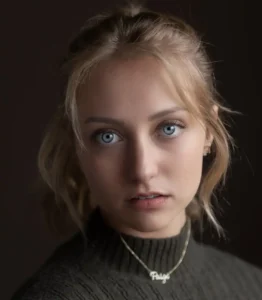 A woman with wavy hair wearing a dark turtleneck and a necklace sits against a dark background, exuding a calm presence.