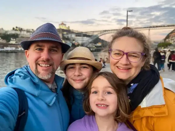 Four people pose for a selfie by the river, with a bridge and hillside city in the background during sunset.