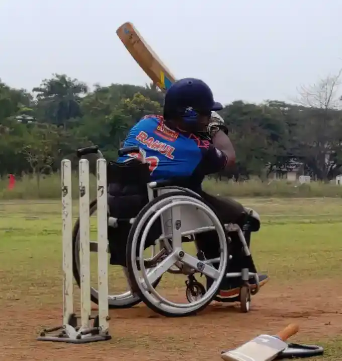 A person in a wheelchair swings a cricket bat toward the stumps on a grassy field, showcasing adaptive sports.