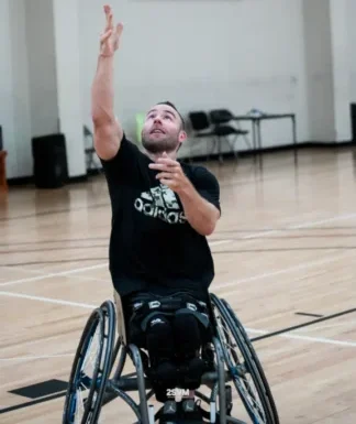 A person in a wheelchair shoots a basketball towards the hoop in a gym, showcasing athleticism and determination.