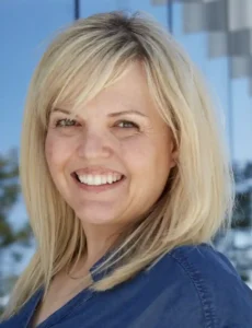 A woman with shoulder-length blonde hair poses in front of a reflective backdrop, styled casually in a blue shirt.
