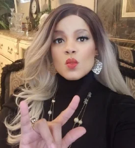 Woman with blonde wig, black turtleneck, and jewelry, making a peace sign.