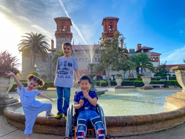 Three children pose in front of a historic building with palm trees, near a fountain, under a clear blue sky at sunset.