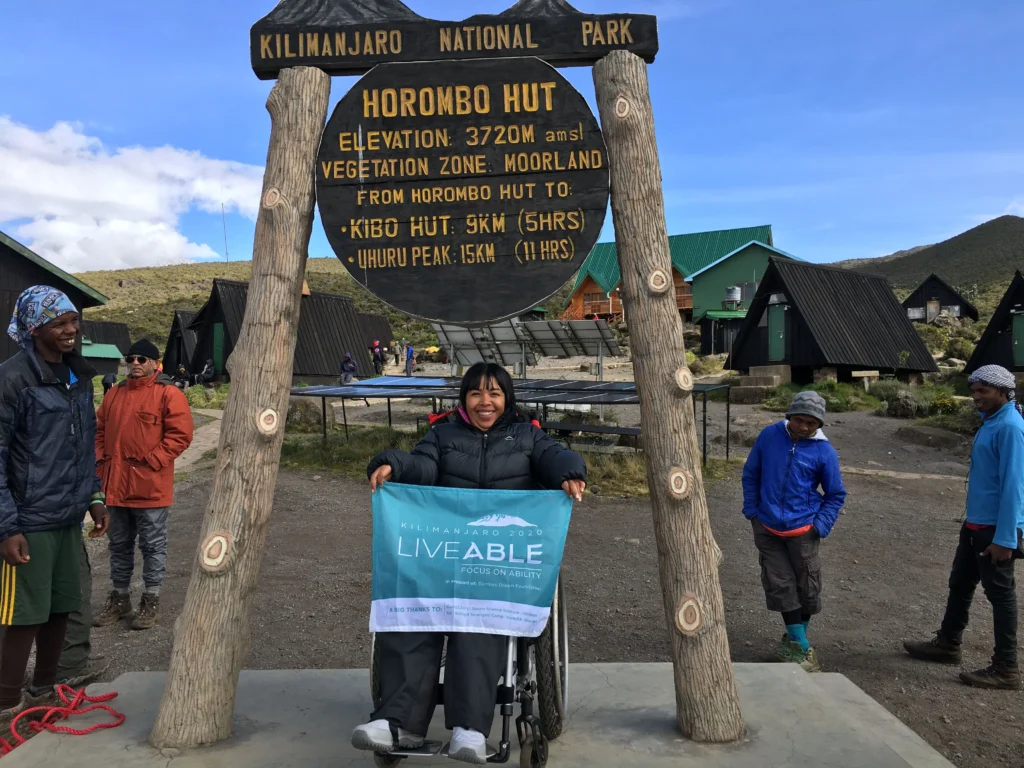 A person in a wheelchair holds a banner at the Horombo Hut sign in Kilimanjaro National Park, surrounded by hikers and cabins.