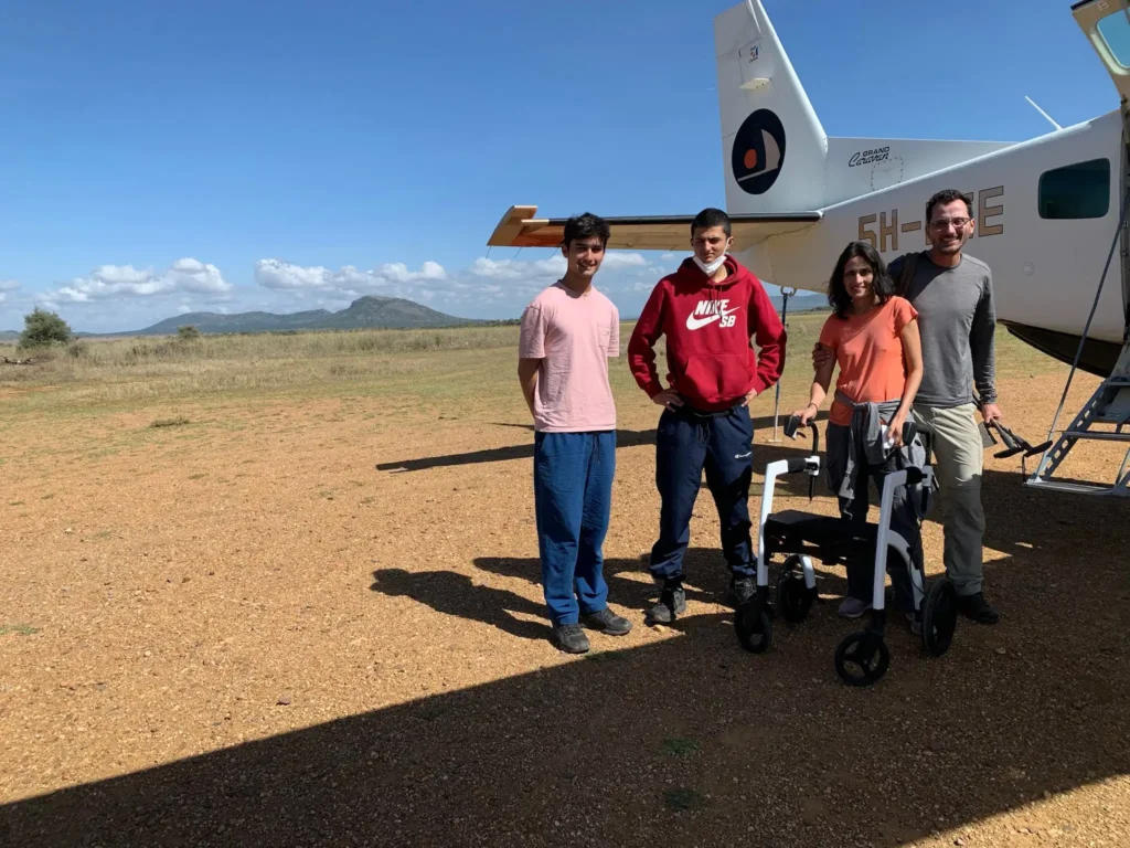A group of four people stands together next to a light aircraft on a dirt airstrip, with mountains and blue sky in the background.