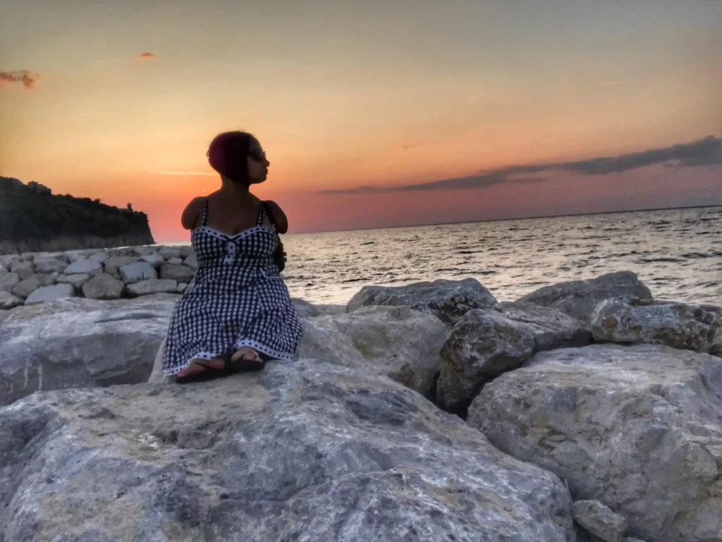 A woman sits on large rocks by the sea, gazing at a colorful sunset over the water, surrounded by nature.
