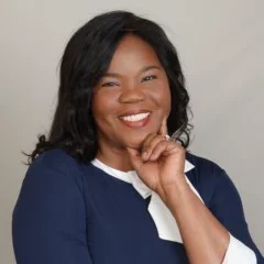 A woman in a navy blue dress with white accents poses thoughtfully, arms crossed, against a plain background.