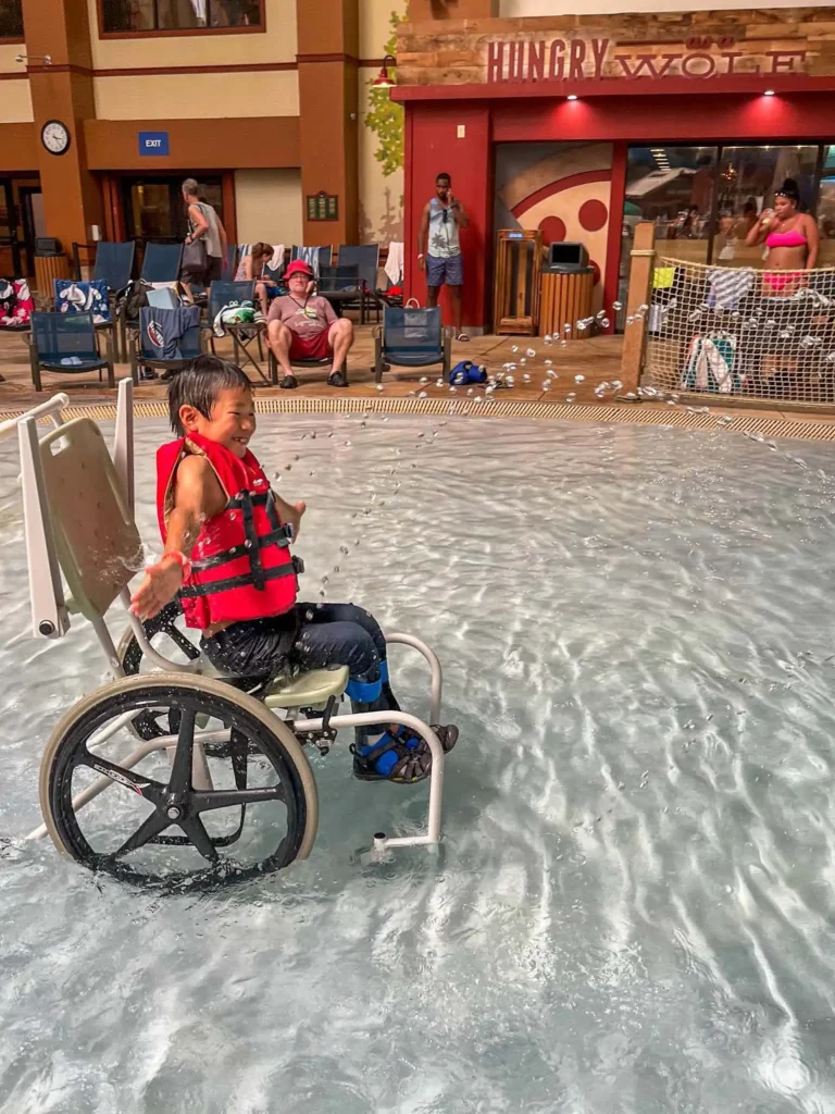 A child in a red life jacket sits in a specialized wheelchair in a pool, surrounded by people enjoying a resort atmosphere.