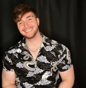 A muscular individual poses against a black backdrop, wearing a black and white patterned short-sleeve shirt and silver chain.
