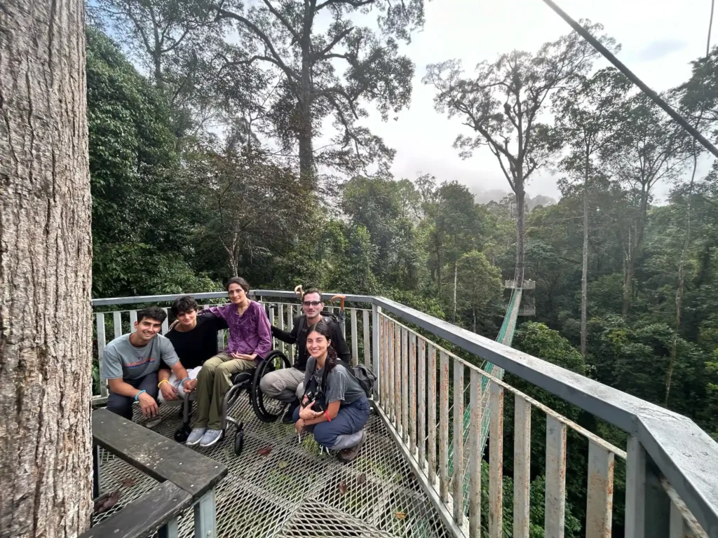 A group of six people poses on a platform surrounded by lush green trees, with a treetop walkway visible in the background.
