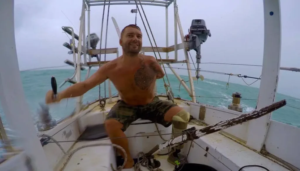 A man with a prosthetic right leg and no left arm sits on a small boat in rough seas under an overcast sky. He is wearing camouflage shorts and a leg brace, and he's smiling as he holds a tool in his hand.