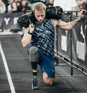 A male athlete with a prosthetic leg is kneeling on a dark floor while holding a heavy sandbag on his shoulder. He is looking down and is gripping a metal barrier with his left hand. The setting appears to be a competition or workout event.