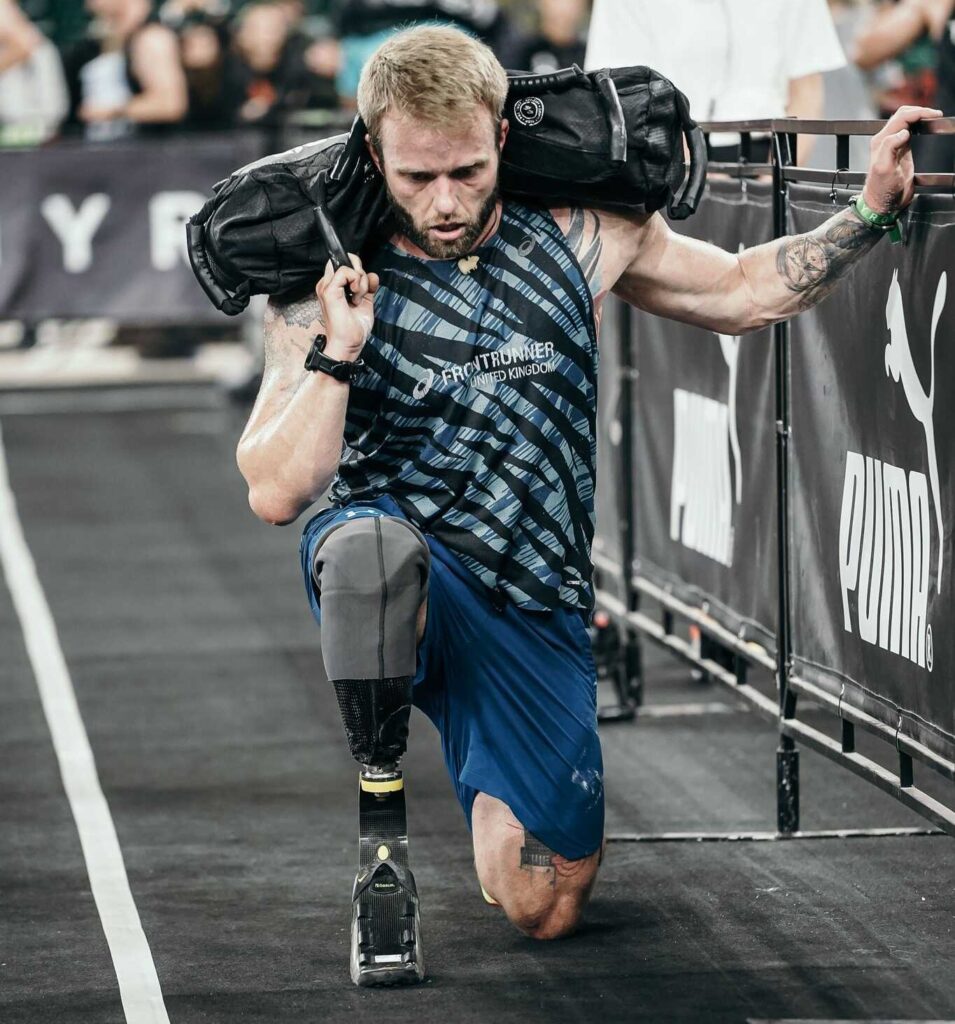 A male athlete with a prosthetic leg is kneeling on a dark floor while holding a heavy sandbag on his shoulder. He is looking down and is gripping a metal barrier with his left hand. The setting appears to be a competition or workout event.