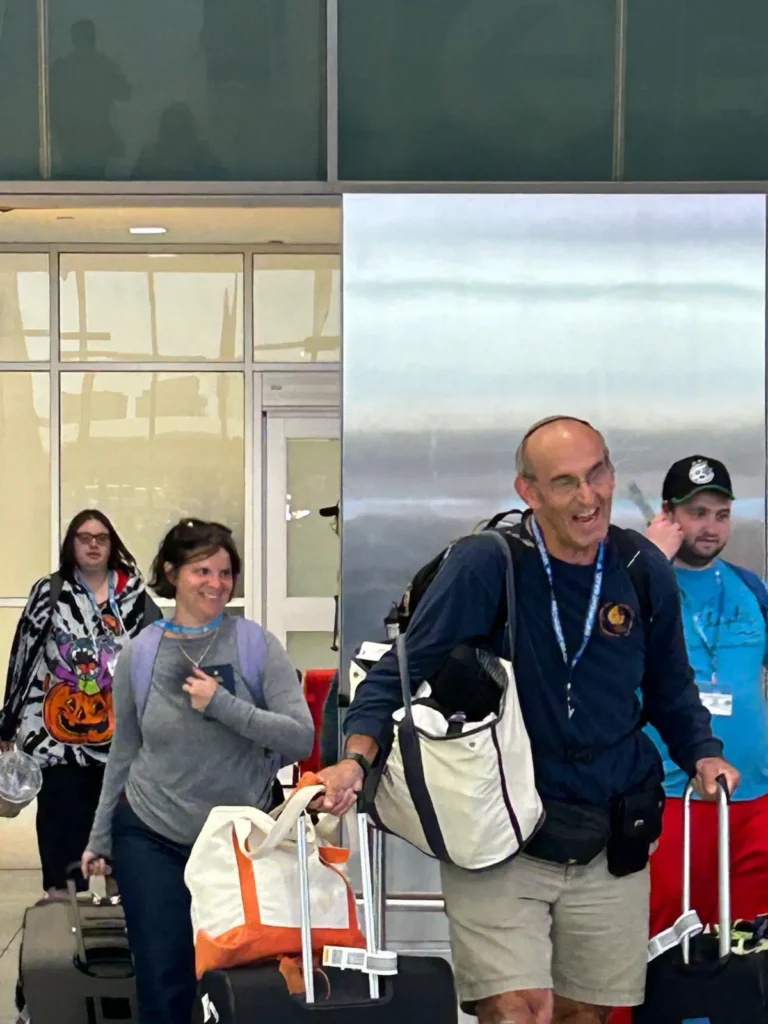A man with a bag slung over his shoulder and another rolling suitcase grins broadly at the camera as he walks with other travelers through an airport.
