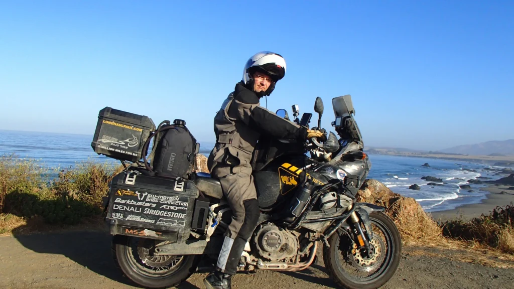 A motorcyclist, fully geared in a helmet, jacket, and pants, sits on a black Yamaha Tenere 700 motorcycle on a dirt road. The bike is equipped with several black bags and panniers. Behind him is a scenic view of the Pacific Ocean coastline under a clear, blue sky.