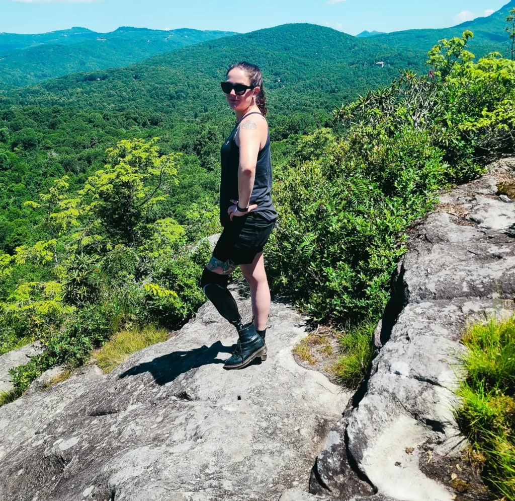 A woman with dark hair and sunglasses stands on a rocky mountain outcrop, looking back at the camera. In the background, there's a vast expanse of lush green mountains and a blue sky with scattered white clouds.