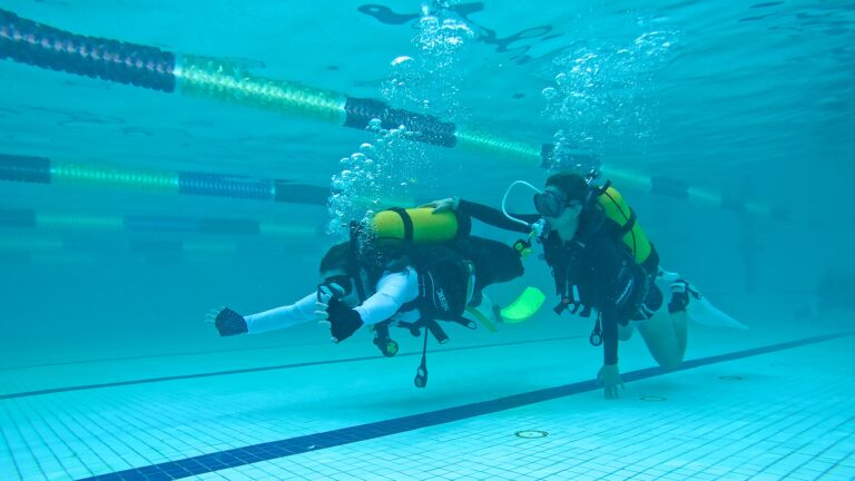 Two people in scuba gear are swimming underwater in a swimming pool. The person on the right is swimming forward, and the person on the left is giving a thumbs-up sign to the camera. Pool lane lines are visible on the surface.