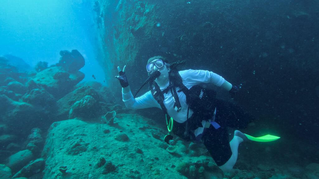 A person in a white long-sleeved shirt and scuba gear swims underwater, giving a peace sign with their left hand. The background is an underwater scene with rock formations and a reef.
