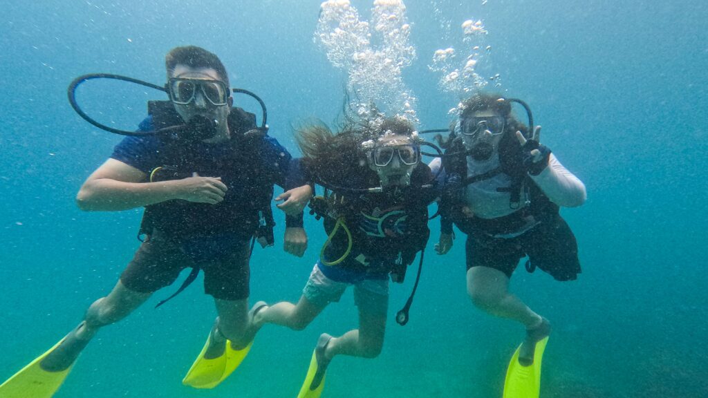 Two men and one woman, all wearing scuba gear, are swimming underwater and posing for a group photo. The person in the middle has long hair and is swimming with a peace sign.