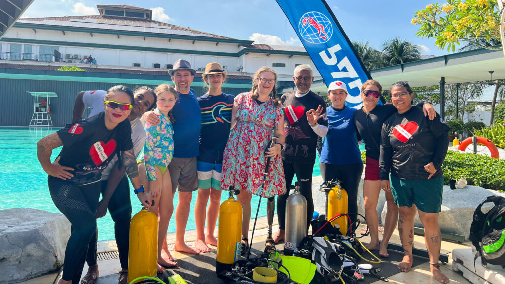 A diverse group of ten people is standing poolside, posing for a group photo. Some are wearing diving gear, and a large blue banner with a logo is visible behind them. Many are smiling and posing with their hands over their hearts, with the hand sign for a heart shape.