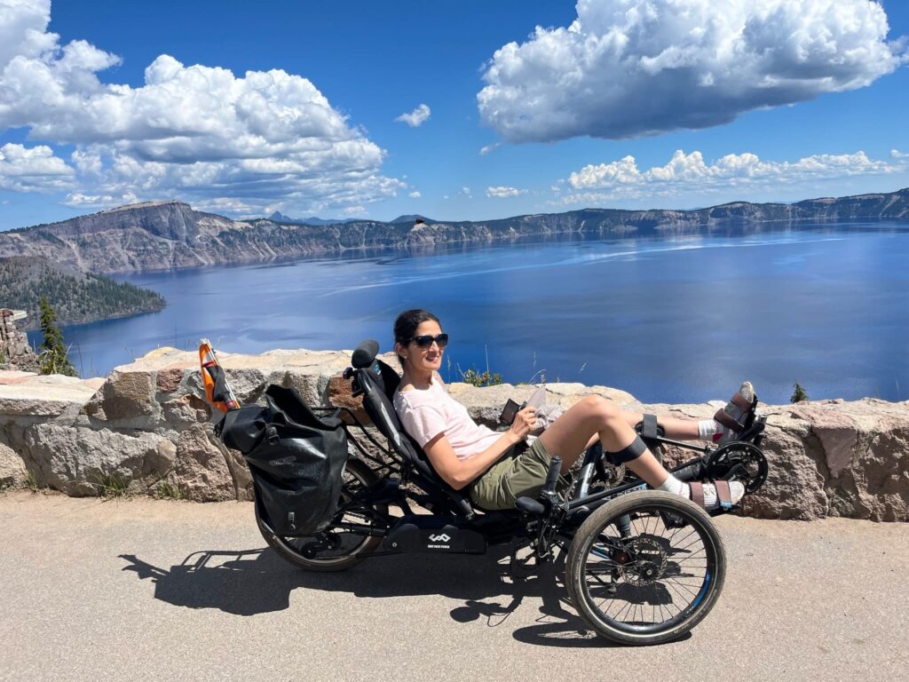 A person sits in a recumbent trike on a paved overlook at Crater Lake. They are smiling and looking at the camera. Behind them, the deep blue lake is surrounded by steep cliffs and forested mountains. The sky is bright blue with large, white clouds.