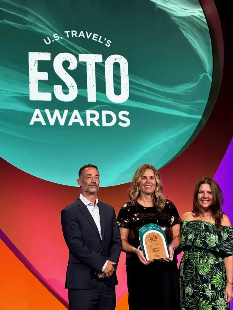 A man and two women stand on a stage in front of a large screen displaying "U.S. TRAVEL'S ESTO AWARDS." The woman in the center is smiling and holding a plaque that reads "All Ways Accessible."