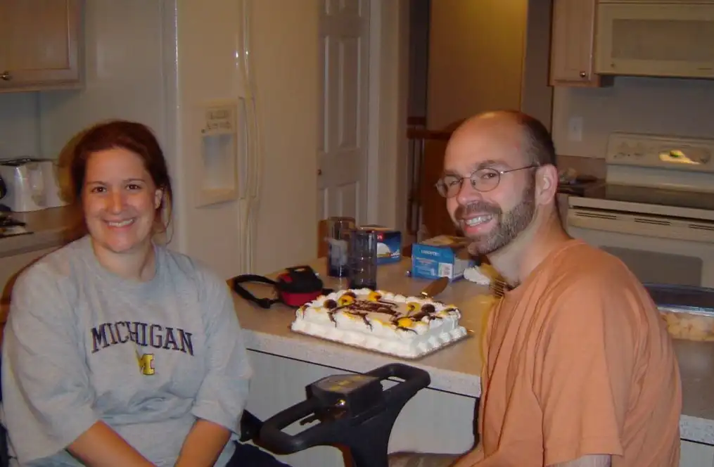 Two people smiling in a kitchen while sitting at a counter with a decorated cake. The woman on the left is wearing a gray University of Michigan sweatshirt, and the man on the right is wearing an orange shirt and glasses. The kitchen has light cabinets, a refrigerator, and a stove in the background.