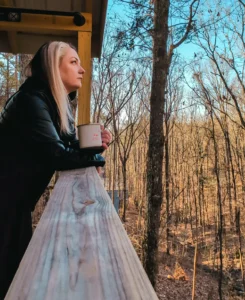 A woman with black and blonde hair stands on a wooden deck, leaning against the railing. She is holding a mug and looking out at a forest of bare trees. The sunlight catches the trees and her face, creating a warm glow.