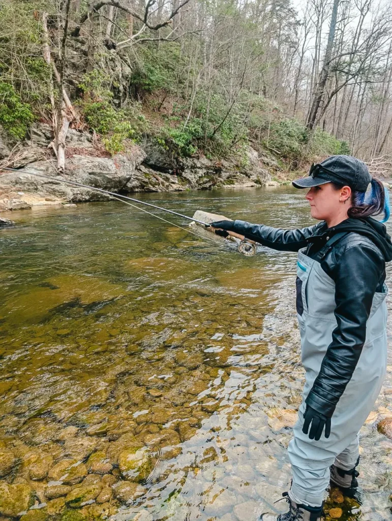 A woman wearing a black jacket, a baseball cap, and chest waders stands in a clear, rocky stream. She is holding a fly-fishing rod and casting a line into the water. The background is a dense forest.