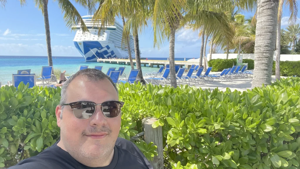 A selfie of a man with a mustache and sunglasses. He is on a sunny beach with palm trees and a large white and blue cruise ship docked in the background.
