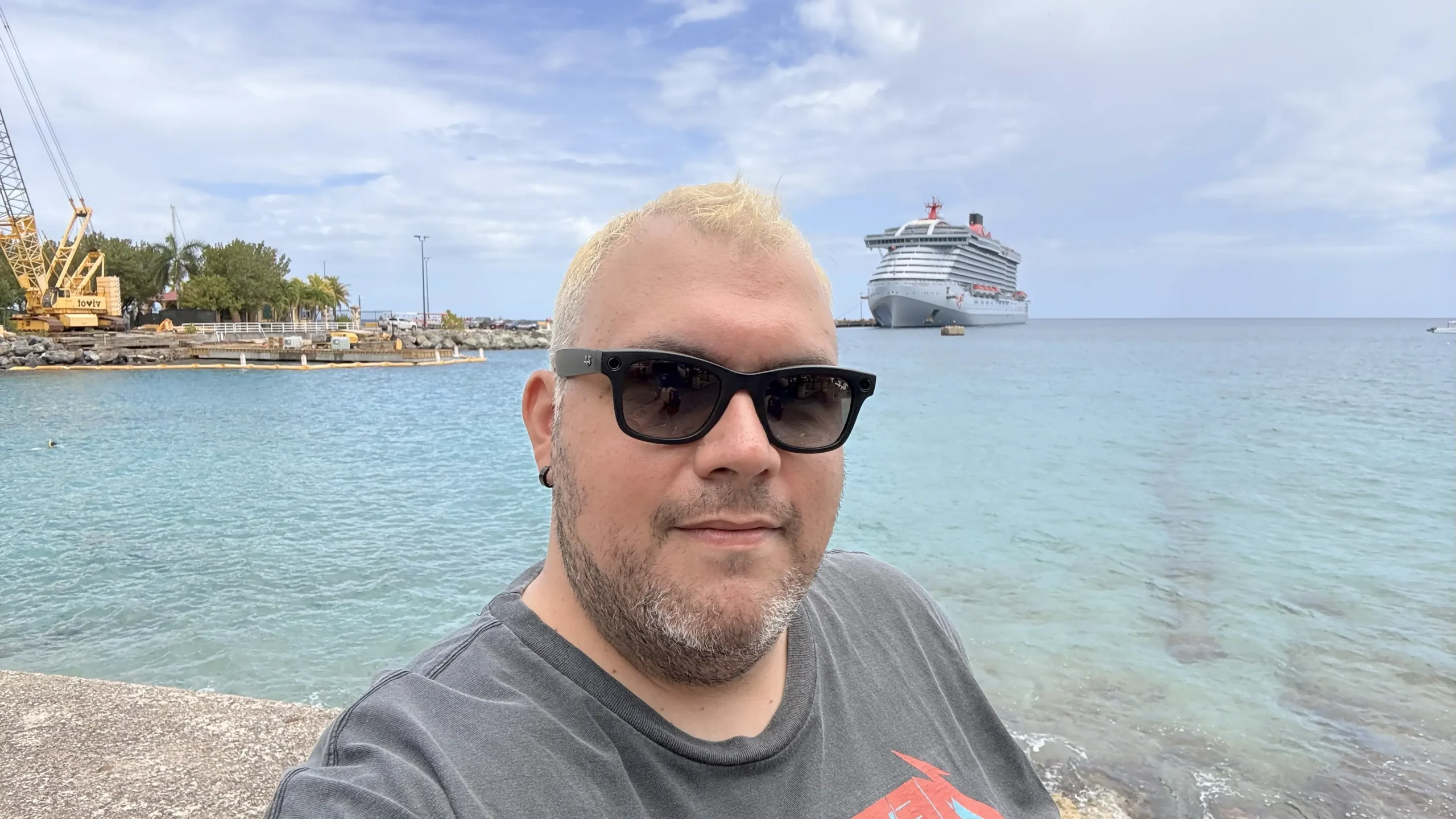 A selfie of a man with short, bleached hair and sunglasses. He's standing by a calm, blue ocean with a large gray and red cruise ship docked in the distance behind him.