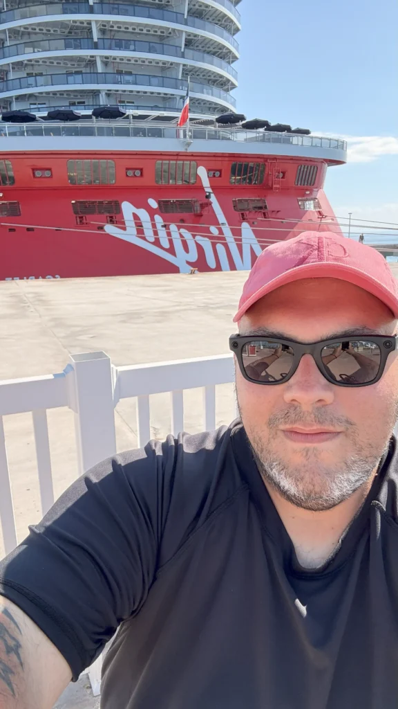 A selfie of a man wearing a red hat and sunglasses. A large red cruise ship with the word 'Virgin' on its side is docked directly behind him.