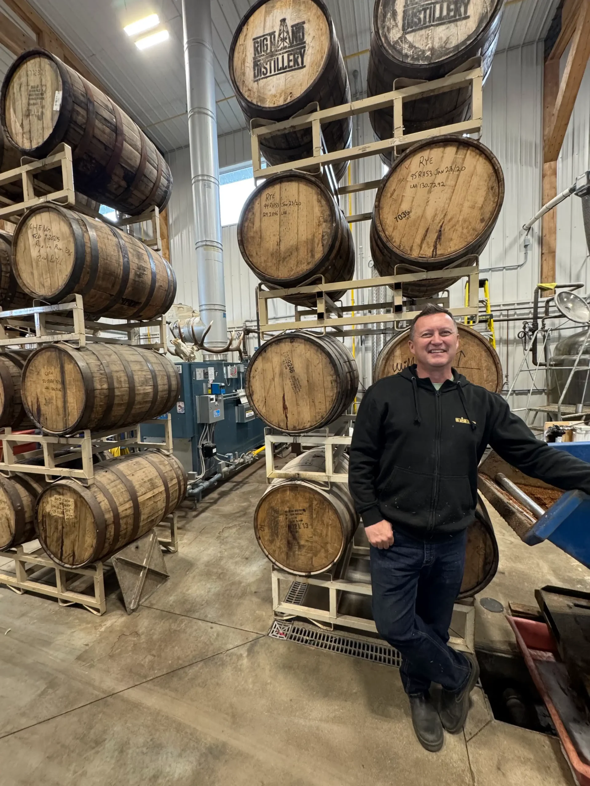 A man in a black hoodie and jeans stands in a distillery, posing next to tall metal racks holding multiple stacked wooden barrels. The barrels have different markings on them.