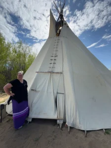 A woman with a black shirt and a long purple skirt stands outside a large, white teepee. The teepee is made of white canvas with a wooden pole structure at the top. The background shows trees and a partly cloudy sky.
