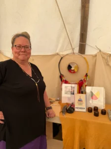 A portrait of a woman with glasses and her hair tied up, standing in a tent or teepee. She is wearing a black shirt and jewelry. A dreamcatcher and some informational charts are displayed on a table behind her.
