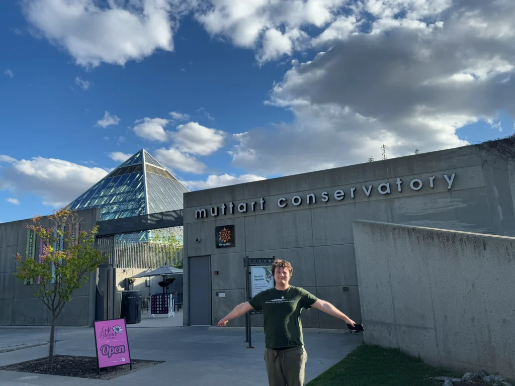 A man with his arms spread out stands in front of the Muttart Conservatory building, a concrete and glass structure with a large pyramid dome on the left. The sky is blue with scattered clouds.