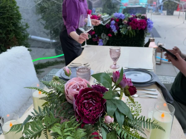 A beautifully set outdoor table for two is visible in the foreground, with a large floral centerpiece of pink and maroon flowers and ferns. Two lit candles in glass holders are on either side. In the background, a server in a purple shirt is attending to a customer in a covered patio area.