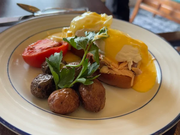 A close-up shot of a plate of Eggs Benedict. The dish includes two poached eggs with hollandaise sauce, a halved tomato, roasted potatoes, and a sprig of parsley on a white plate with a blue rim.