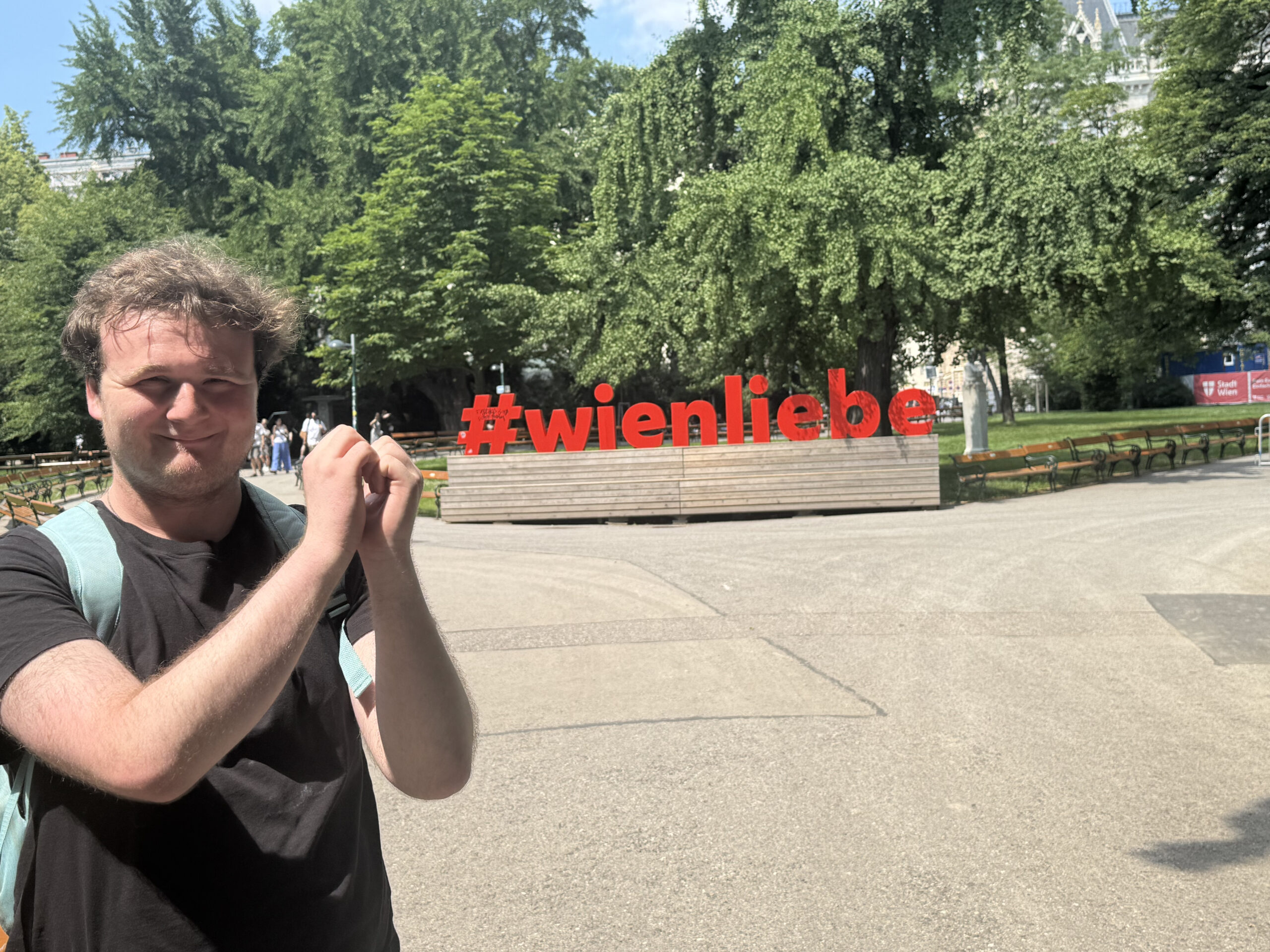 A young man with light brown, wavy hair smiles at the camera, holding his hands clasped together near his chest. He's wearing a black T-shirt and a light blue backpack. In the background, a large, modern art installation spells out "#wienliebe" in red letters on a wooden base, surrounded by green trees and park benches under a clear, blue sky.