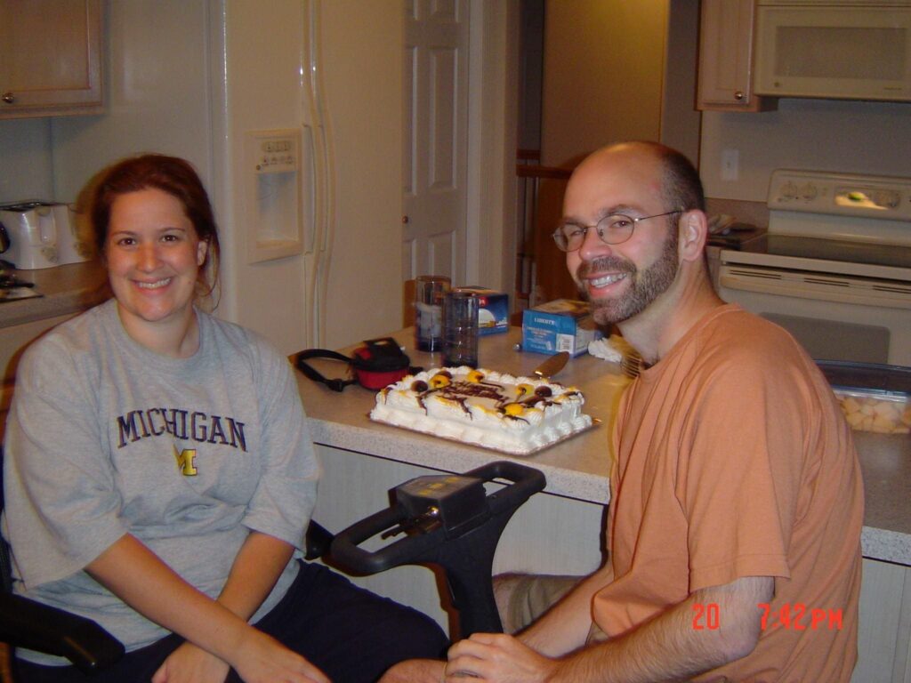 A smiling woman with brown hair sits in a mobility scooter at a kitchen counter, wearing a gray sweatshirt with "MICHIGAN" and a logo on it. A smiling man with a beard and glasses sits next to her. A cake with suns, clouds, and a palm tree is on the counter between them.