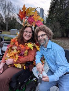 A smiling woman sits in a wheelchair outdoors, wearing a costume made of autumn leaves. A man with a beard and glasses sits next to her, wearing a light blue button-down shirt and a Bob Ross-style wig. He is holding a painter's palette and a paintbrush.