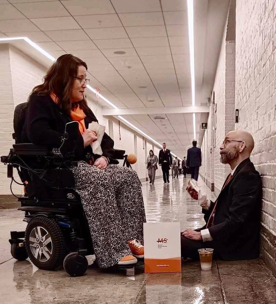 A woman in a black jacket and long patterned skirt sits in a wheelchair in a hallway. She is holding a white bag and talking to a man in a suit who is sitting on the floor leaning against the wall, holding a white bag and a coffee cup. People are walking in the background of the long hallway.