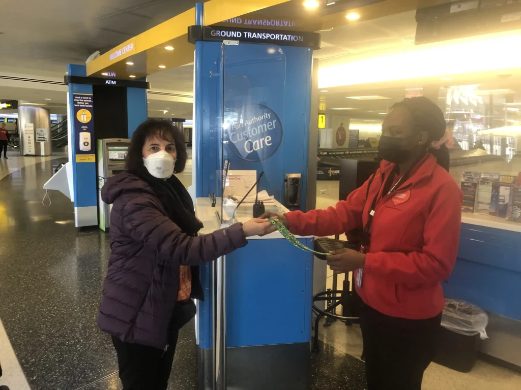 A woman wearing a face mask and a purple jacket receives a lanyard from an airport customer care employee at a Port Authority desk.