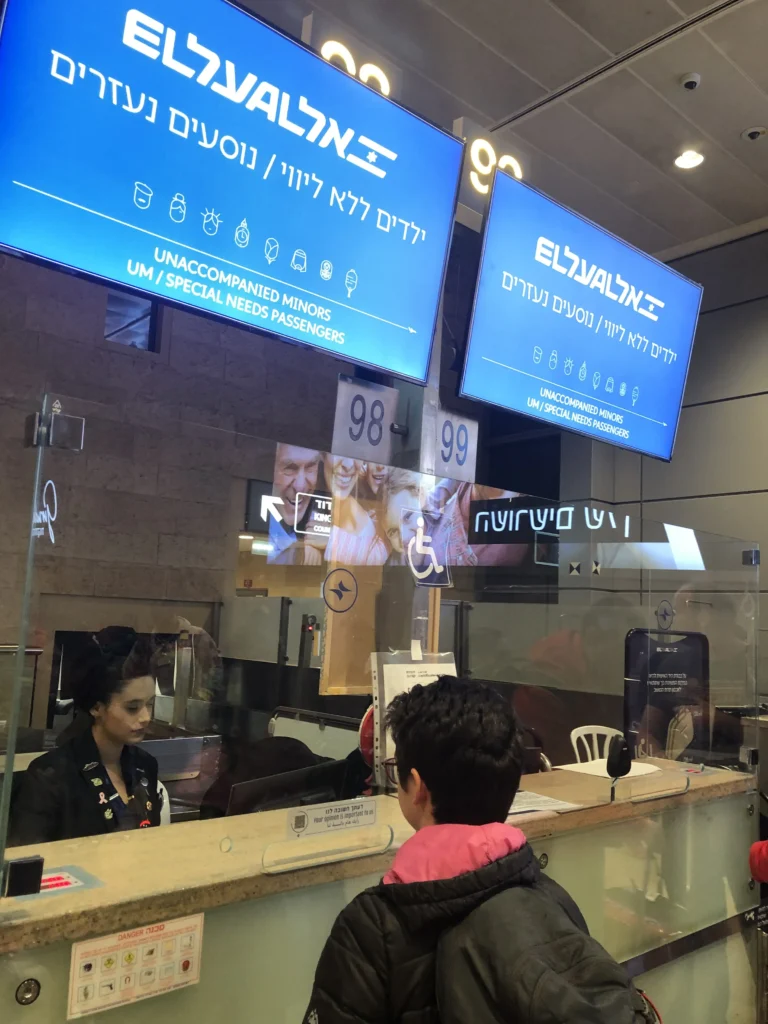 A young person with their back to the camera stands at a check-in counter for El Al airlines, with signs overhead in both Hebrew and English for "Unaccompanied Minors / Special Needs Passengers." An airline employee is seated at the desk, looking at the traveler.