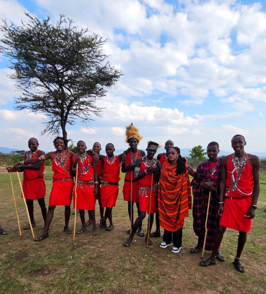 A group of people, including ten men dressed in red Maasai attire and one woman in a striped shawl, pose for a group photo in a grassy field with a large tree behind them. They are smiling, and some are holding wooden staffs. The background shows rolling hills and a partly cloudy sky