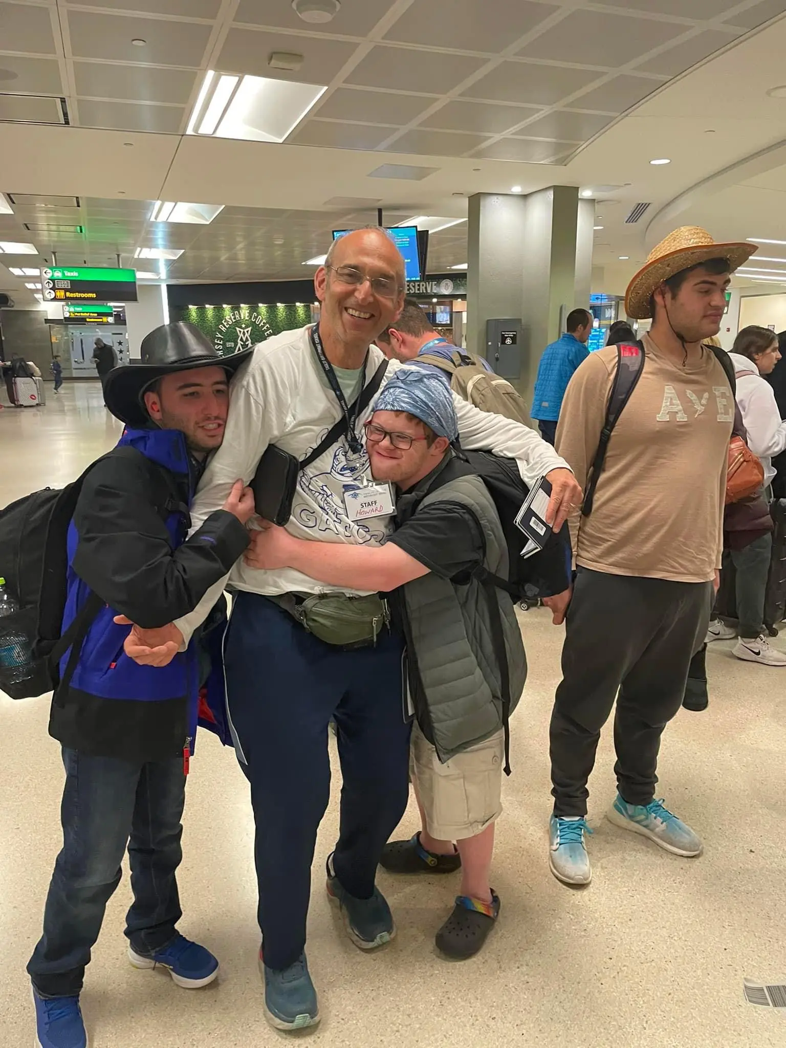 A group of three young men and an older man stand together in an airport. The older man is laughing while hugging one of the younger men, who is wearing a hat, a fanny pack, and glasses.