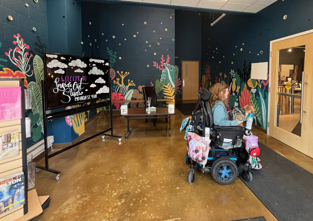 The interior of a bright and colorful art studio. The walls are painted a dark teal with a mural of whimsical, stylized plants and flowers. A large television on a stand to the left displays a "Welcome to InsideOut Studio" message. A woman in a motorized wheelchair sits near a table, facing away from the camera.